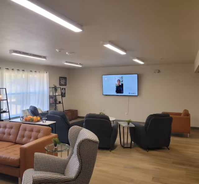 A cozy living room area in an assisted living facility with multiple comfortable chairs and sofas arranged around small tables. A flat-screen TV is mounted on the wall displaying a person’s image. The room has large windows with sheer curtains allowing natural light to enter, and the floor is wooden.
