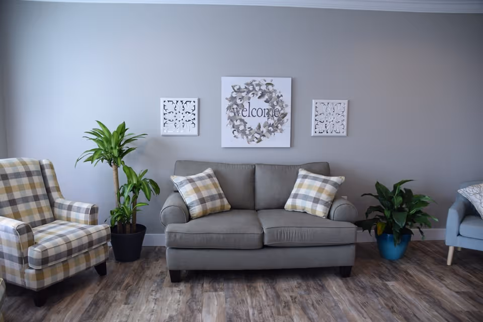 A cozy living room area with a gray sofa adorned with two plaid pillows, a matching plaid armchair to the left, and a light blue armchair partially visible on the right. There are two potted plants on either side of the sofa, and three wall decorations above it, including a central piece with a floral wreath and the word 'welcome'. The room has light gray walls and wood-patterned flooring.