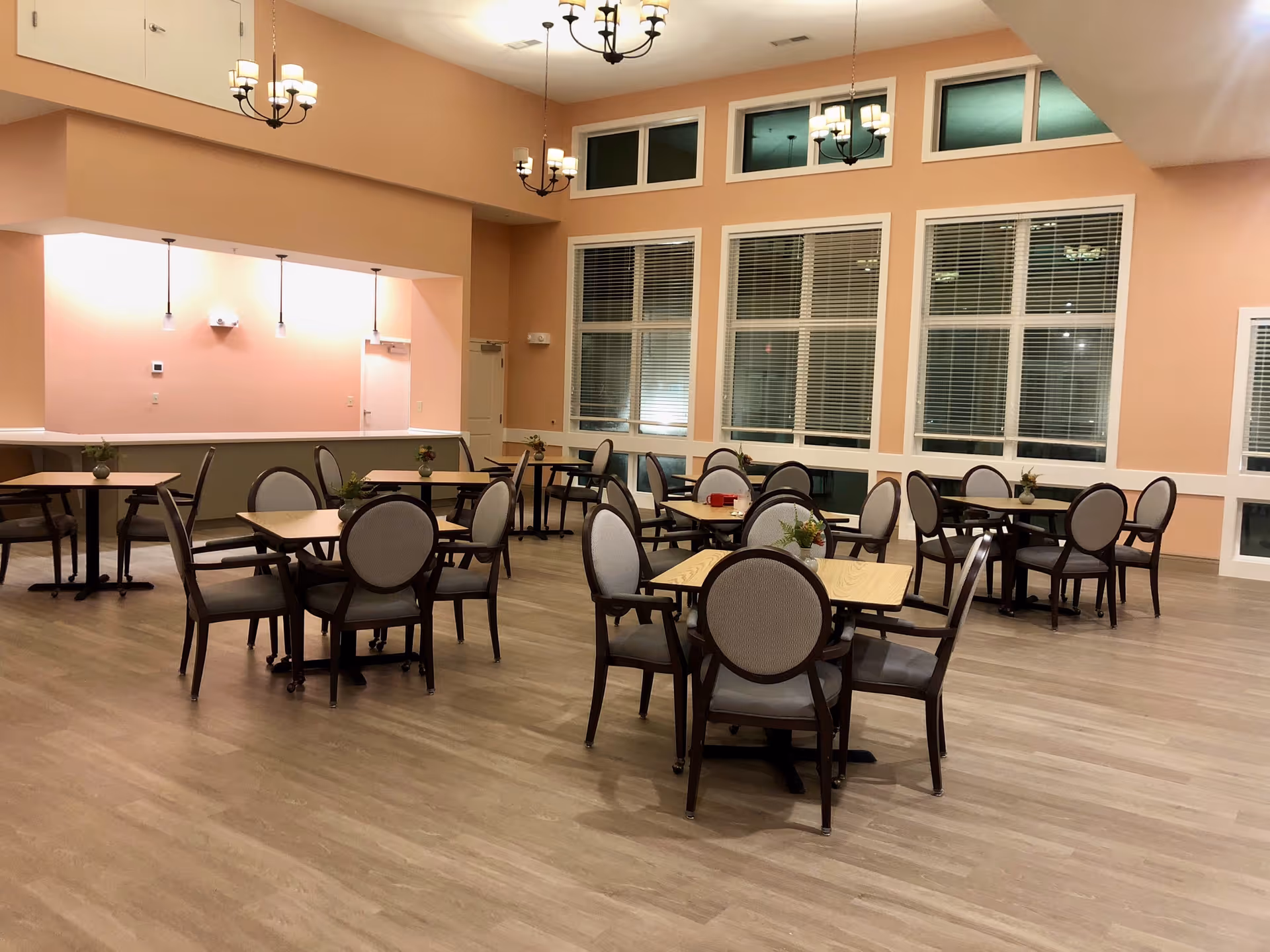 A spacious dining room with several wooden tables and cushioned chairs arranged neatly. The room has large windows with blinds, peach-colored walls, and multiple chandeliers hanging from the ceiling. Small plants are placed on each table, and the floor is covered with light wood flooring.