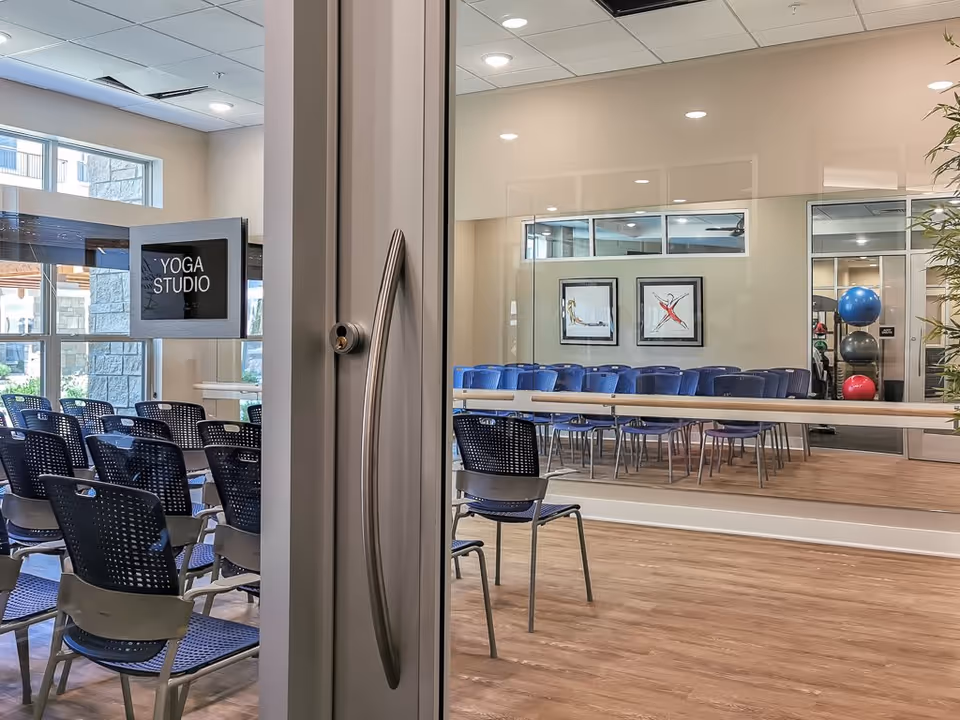 Interior view of a yoga studio with rows of blue chairs, a mirrored wall, and exercise balls visible through a glass door labeled "YOGA STUDIO".
