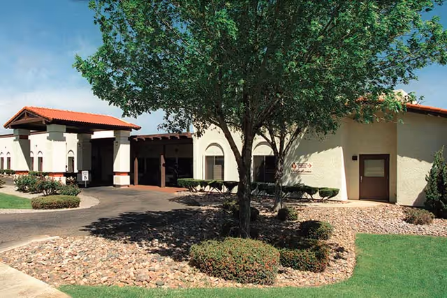 Exterior view of Life Care Center of Sierra Vista showing a single-story building with beige walls and a red-tiled roof. There is a driveway leading to a covered entrance, surrounded by landscaped areas with rocks, bushes, and a large tree in the foreground under a clear blue sky.