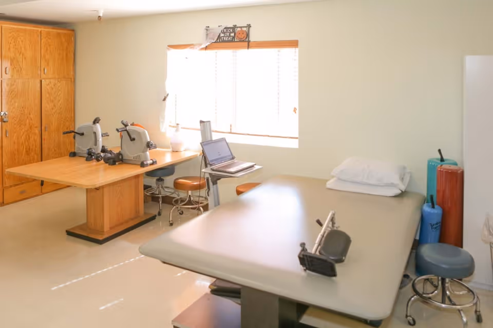 A bright rehabilitation therapy room with a treatment table, exercise equipment on a wooden table, stools, cabinets, and a laptop by the window.