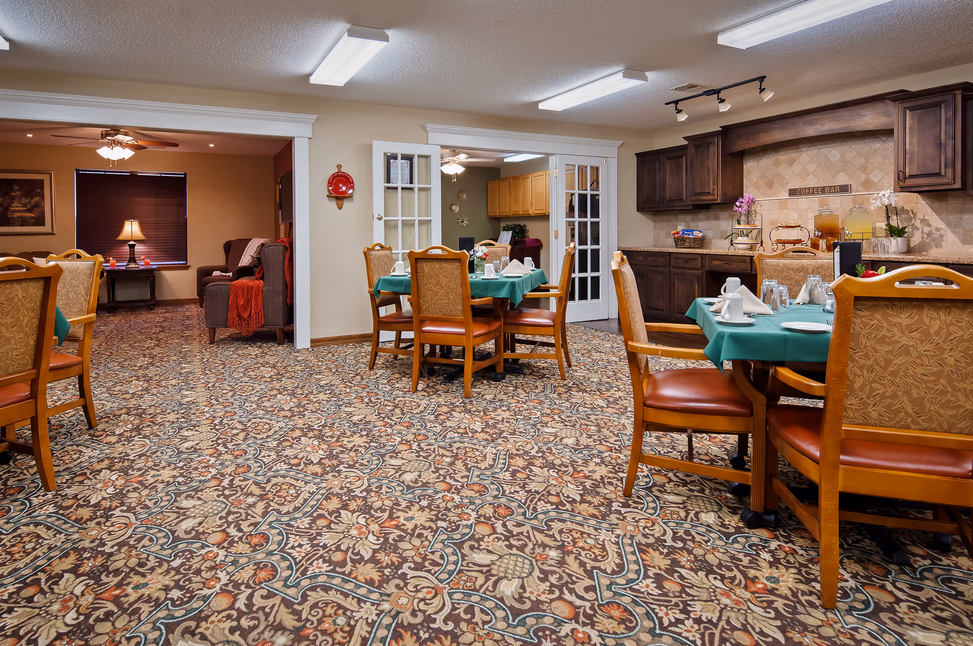 Dining room with several set tables, patterned carpet, a kitchenette area, and a seating lounge in the background.