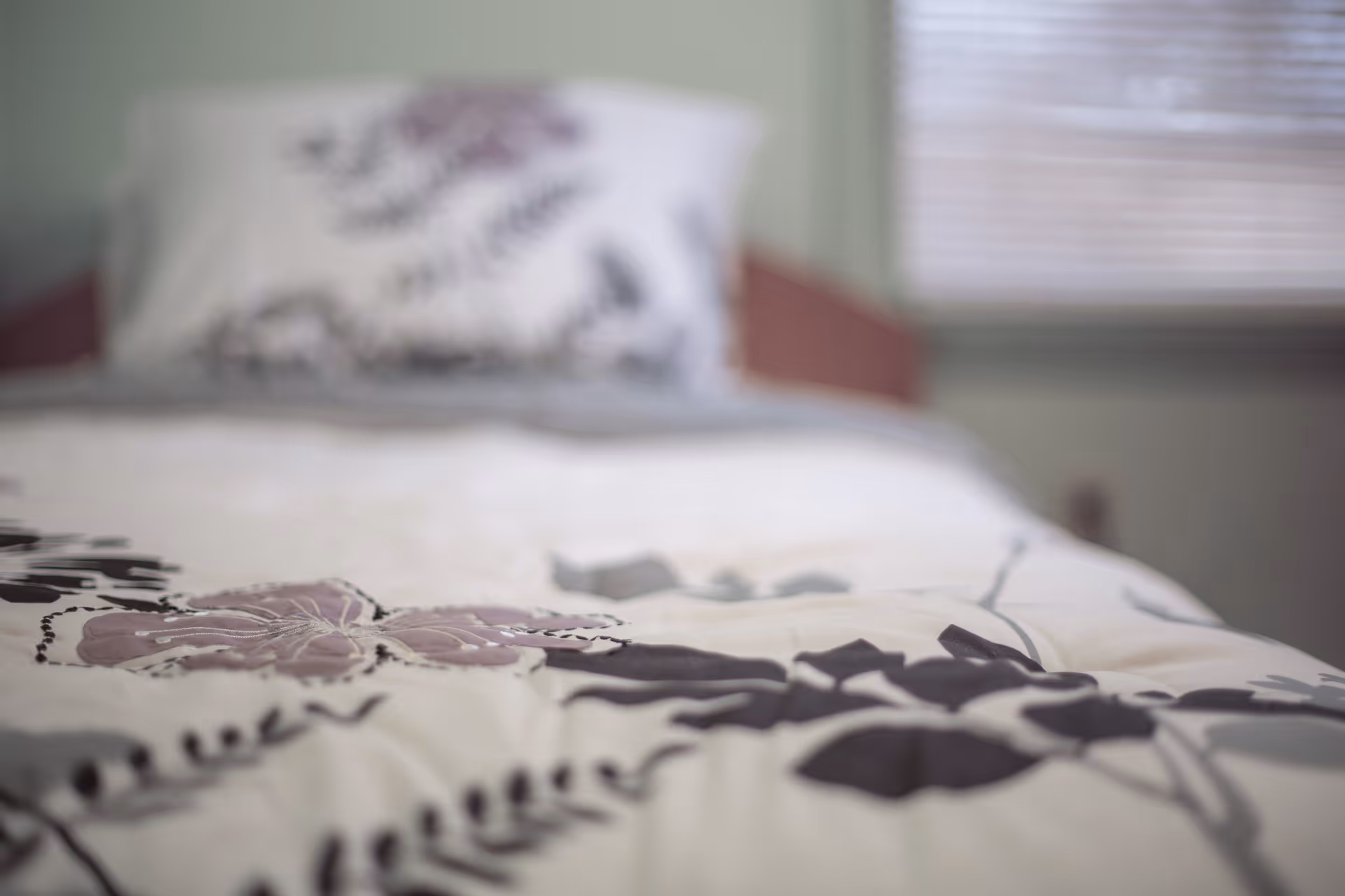Close-up view of a bed with a floral patterned comforter and a matching pillow in a softly lit bedroom with a window in the background.