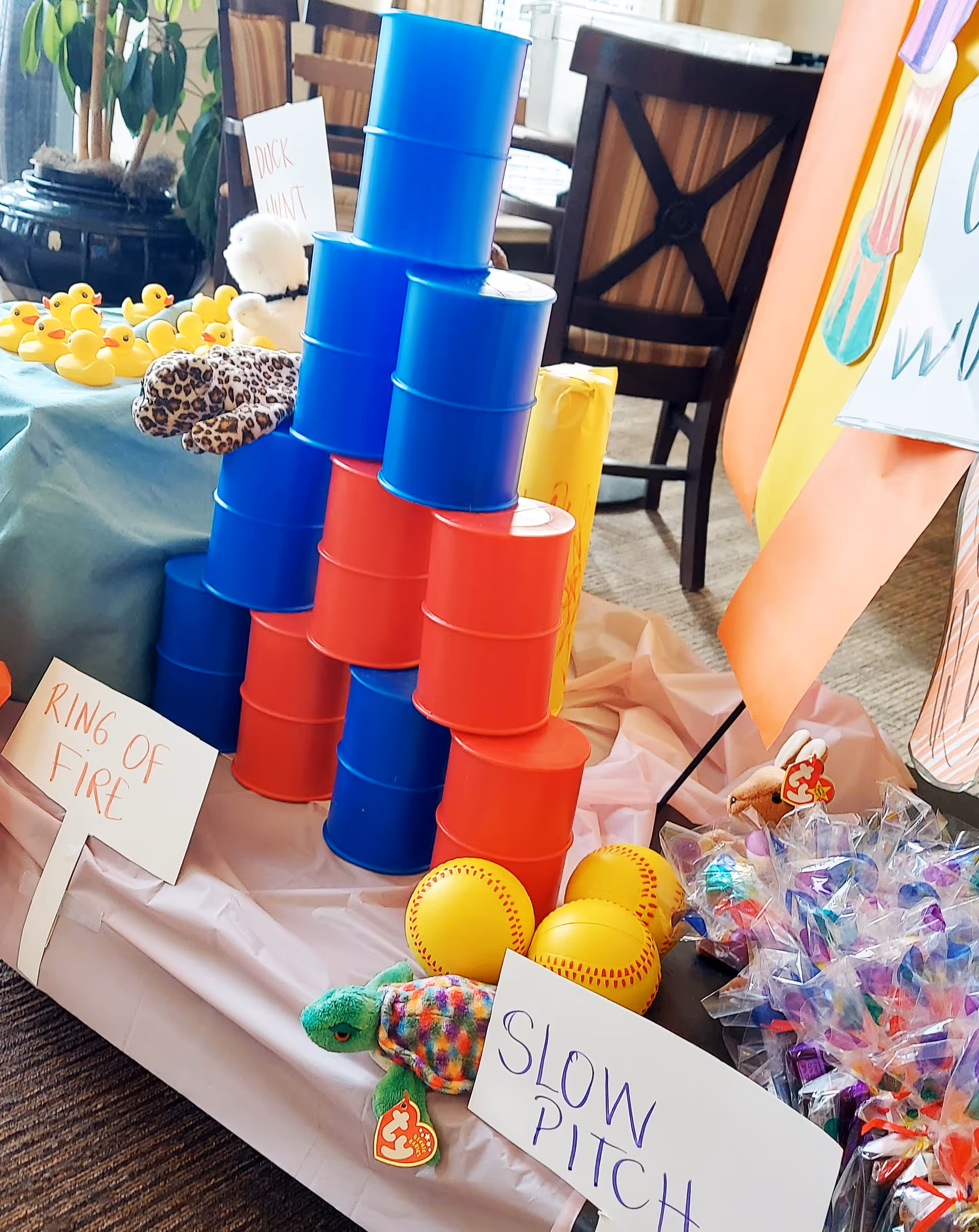 A table set up with colorful game props including stacked red and blue cans, yellow softballs, a plush turtle, and a collection of small rubber ducks. Handwritten signs label the games as 'Ring of Fire,' 'Slow Pitch,' and 'Duck Hunt.' The setting appears to be an indoor common area with chairs and a plant in the background.