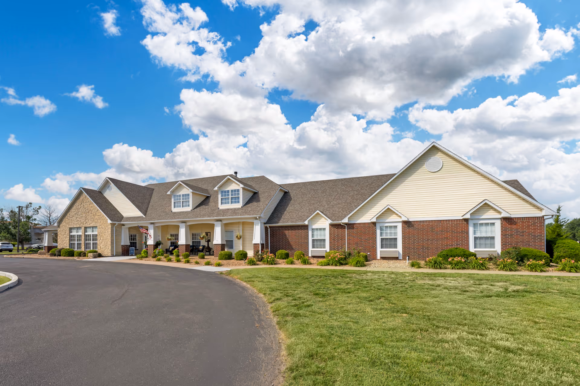 Exterior view of a single-story senior living facility building with a combination of brick and siding walls, a covered porch with seating, well-maintained landscaping, and a curved driveway under a partly cloudy blue sky.