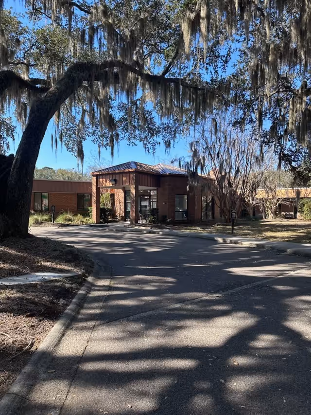 Exterior view of a single-story brick building with a covered entrance, surrounded by trees with hanging Spanish moss and a clear blue sky.