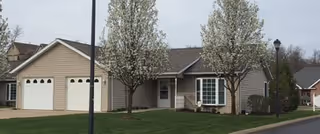 Single-story beige residential building with an attached two-car garage, small front lawn, and two flowering trees.