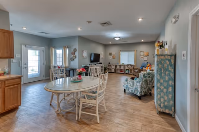 A spacious senior living common area with light blue walls and wood flooring. The room features a round dining table with four chairs, a patterned armchair, a TV mounted on the wall, storage cubbies, and a cabinet with floral design. There are windows with curtains and a glass door leading outside, allowing natural light to fill the room.