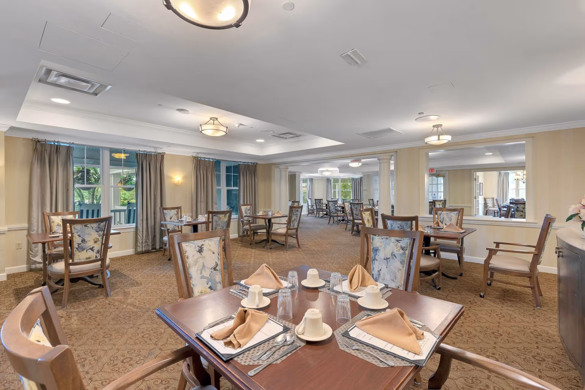 A spacious dining room with multiple wooden tables and chairs arranged neatly. Each table is set with beige napkins, white cups, glasses, and silverware. The room has beige walls, large windows with curtains, carpeted floors, and ceiling lights providing bright illumination. There is a large mirror on one wall reflecting the room.