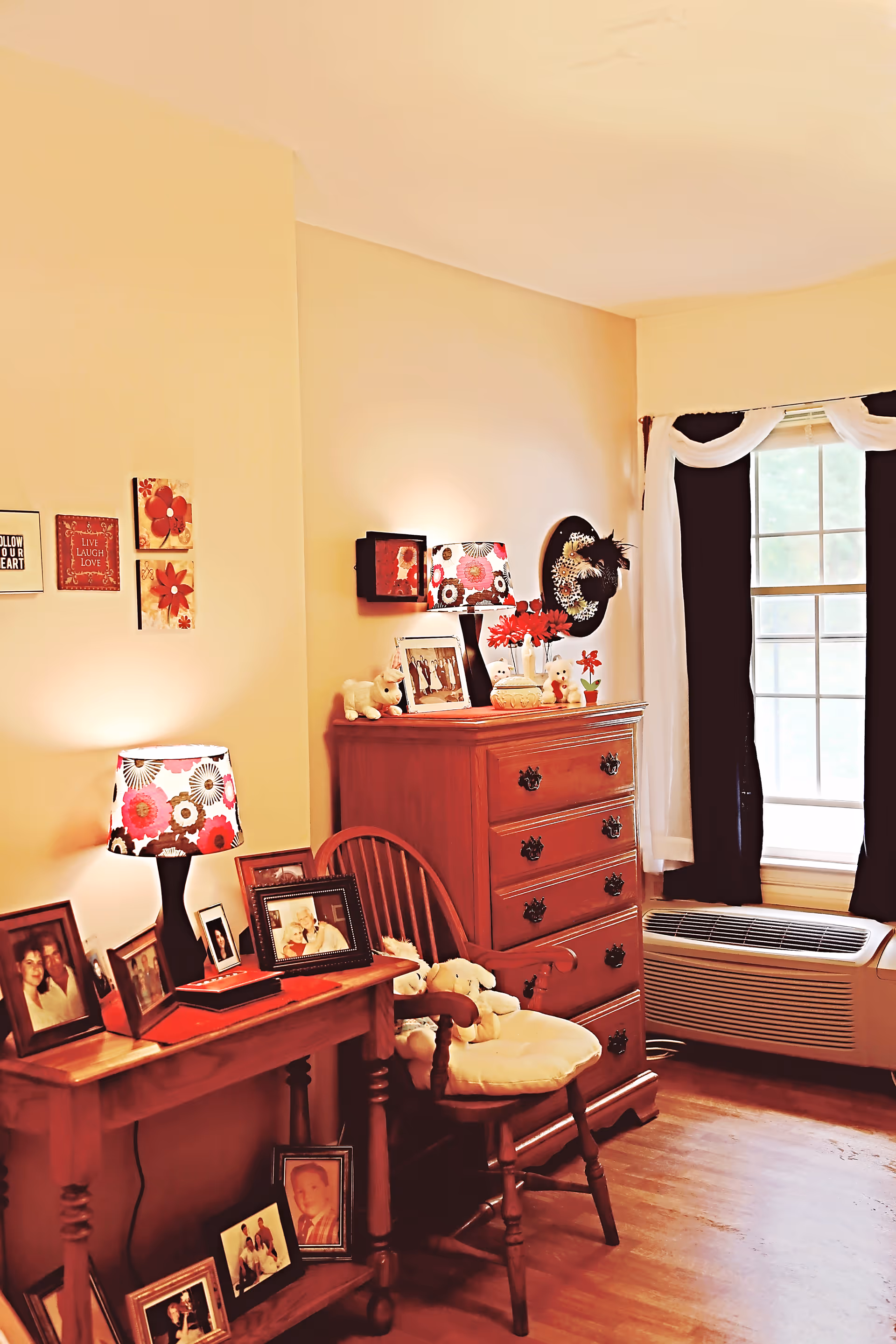 A cozy senior living bedroom corner featuring a wooden dresser with decorative items and framed photos on top, a wooden chair with a cushion and stuffed animals, a wooden side table with a floral lampshade and multiple framed photos, light yellow walls adorned with small wall art, and a window with black and white curtains letting in natural light.