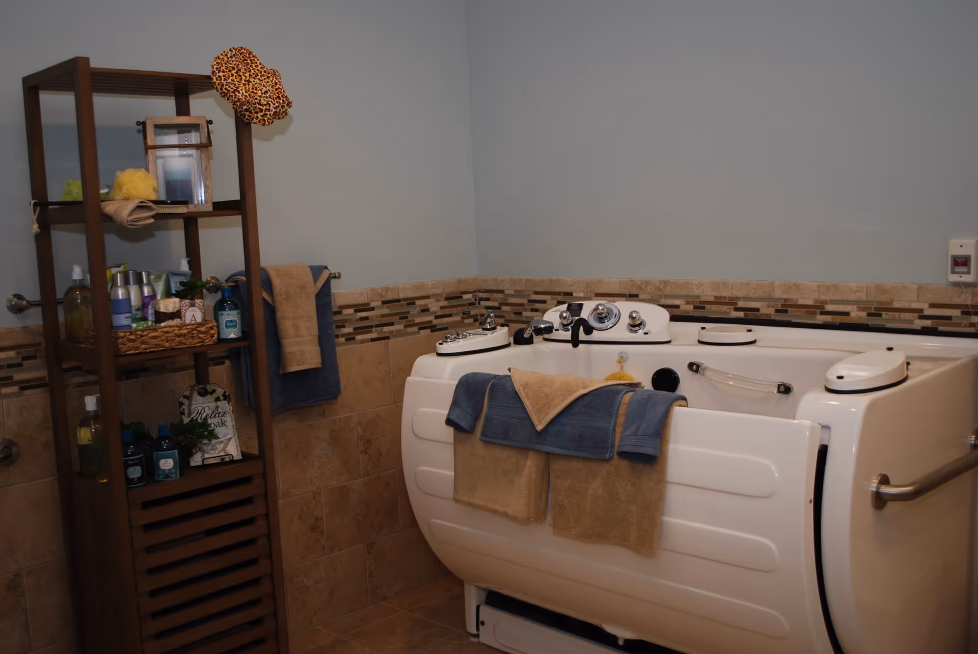 A bathroom featuring a large white walk-in bathtub with control knobs and handrails. The tub has blue and beige towels hanging over the side. To the left, there is a wooden shelving unit holding various bath products and towels. The walls are light blue with a decorative tile border, and the floor is tiled in a beige color.