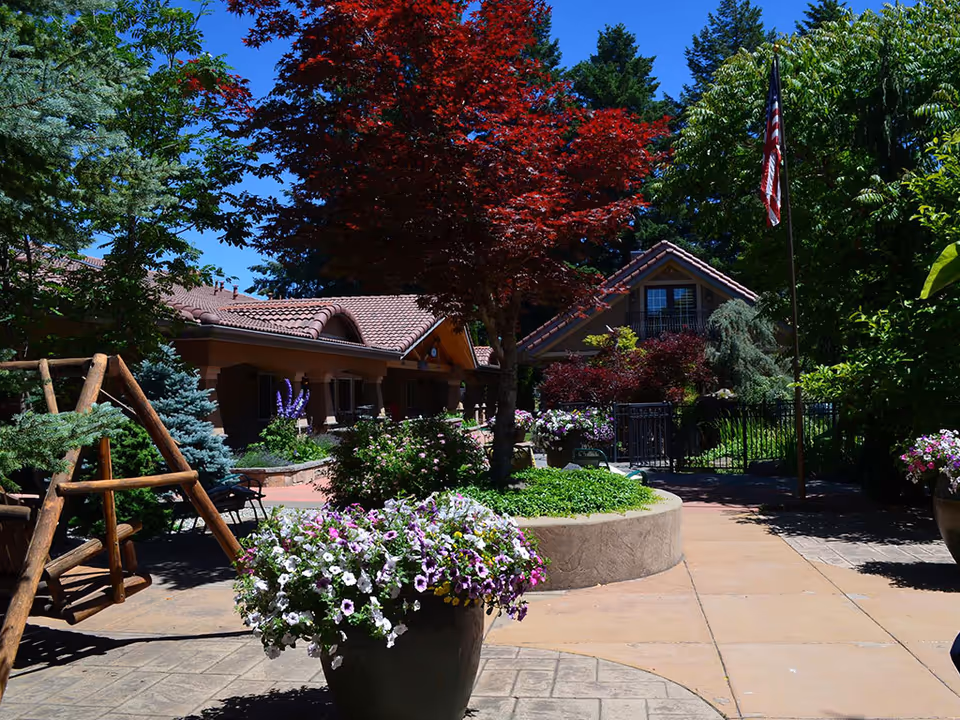 Outdoor courtyard area with a wooden swing, large flower pots filled with colorful flowers, a variety of trees including a red-leafed tree, and a building with a tiled roof in the background under a clear blue sky.