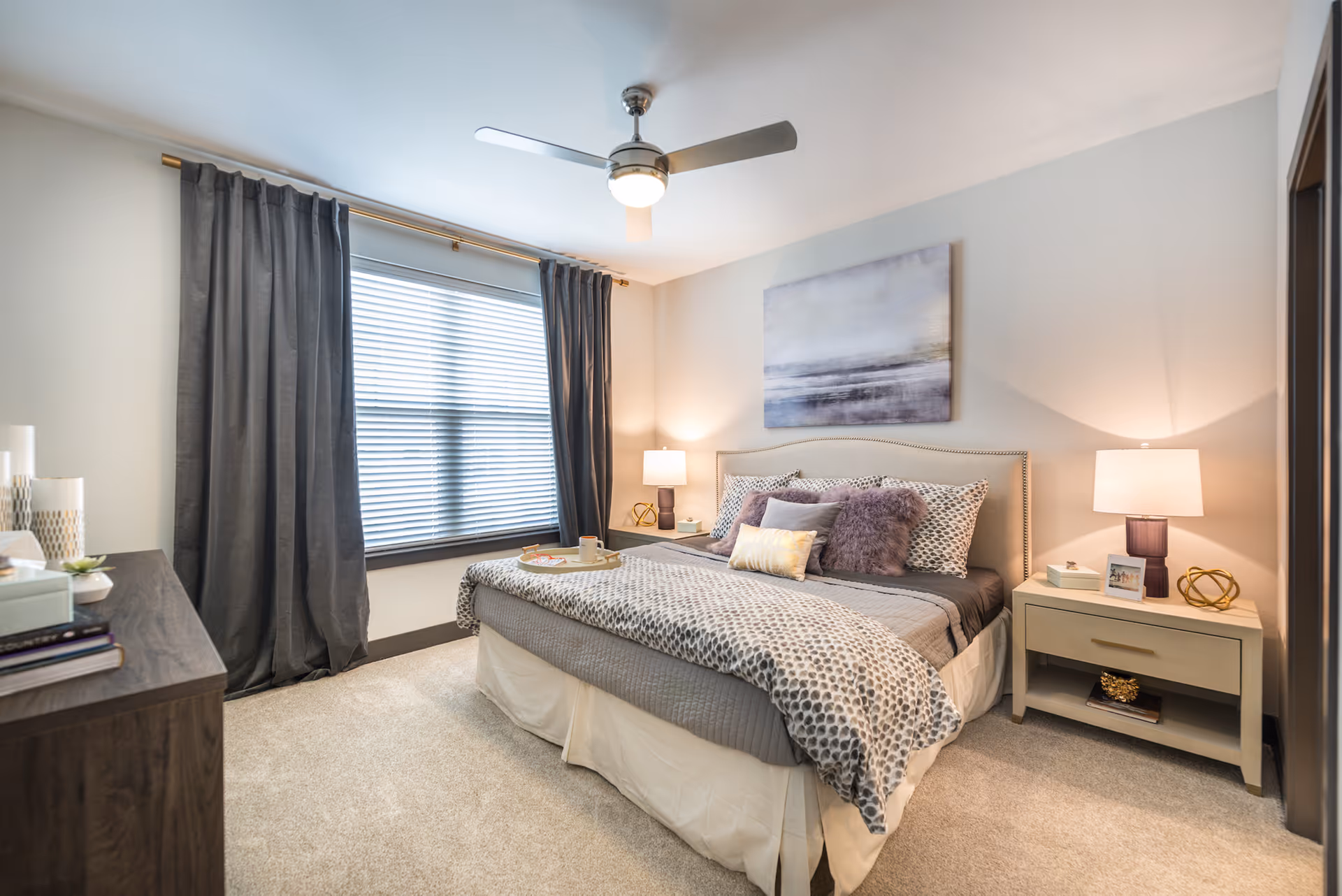 A cozy bedroom with a large bed featuring patterned bedding and multiple pillows. There are two nightstands on either side of the bed, each with a lamp and decorative items. A large window with dark curtains is on one wall, and a ceiling fan with a light is mounted on the ceiling. The room has light-colored walls and carpeted flooring.