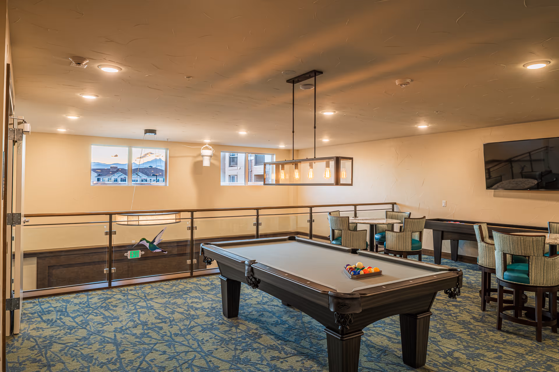 A well-lit recreational room with a pool table in the center, surrounded by several tables and chairs. The room has large windows letting in natural light, a modern hanging light fixture above the pool table, and a flat-screen TV mounted on the wall. The floor is covered with a patterned carpet, and there is a glass railing overlooking a lower level.