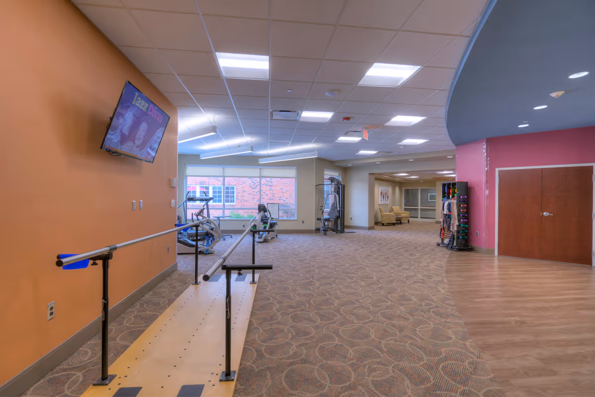 Interior view of a rehabilitation or exercise area in a senior living facility with parallel bars for walking practice, exercise equipment near large windows, a TV mounted on an orange wall, and a seating area in the background.