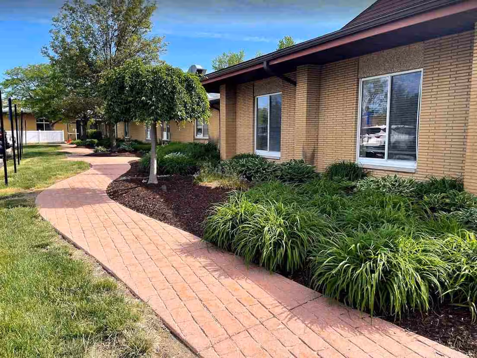 Curved brick walkway bordered by green grass and landscaped plants leading alongside a single-story brick building with windows under a clear blue sky.