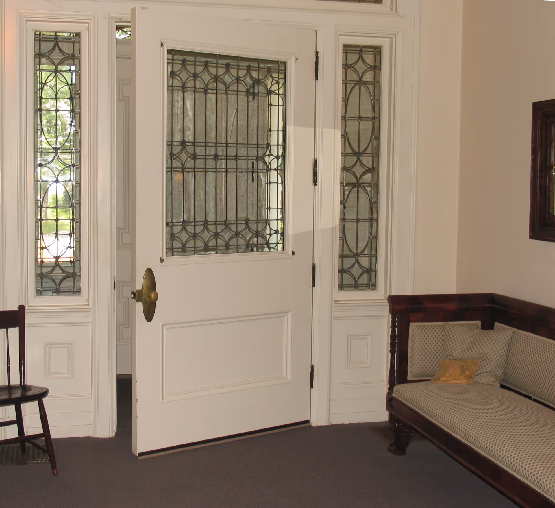An interior view of a room with a partially open white door featuring decorative stained glass panels. To the right is a wooden bench with patterned cushions, and to the left is a wooden chair. The walls are light-colored, and there is a framed mirror or picture on the right wall.