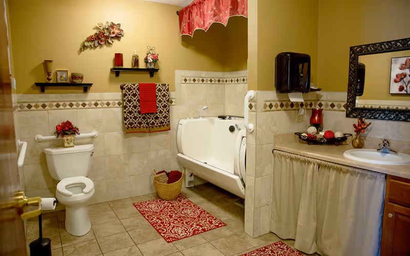 A well-decorated bathroom with beige tiled walls and floor. It features a white toilet with a red flower arrangement on top, a walk-in bathtub with grab bars, a sink with a large ornate mirror above it, and red and brown patterned towels hanging on the wall. There are decorative shelves with small items and a red curtain valance above the bathtub. Red patterned rugs are placed on the floor.
