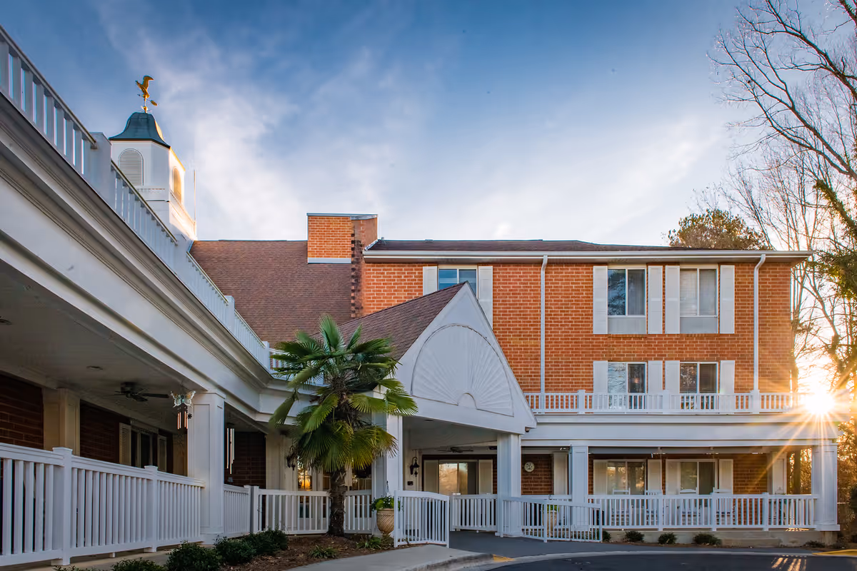 Exterior view of a brick senior living facility building with white railings and a covered entrance. The building has multiple windows with white shutters, a cupola with a weather vane on the roof, and a palm tree near the entrance. The sun is setting in the background, casting a warm light.