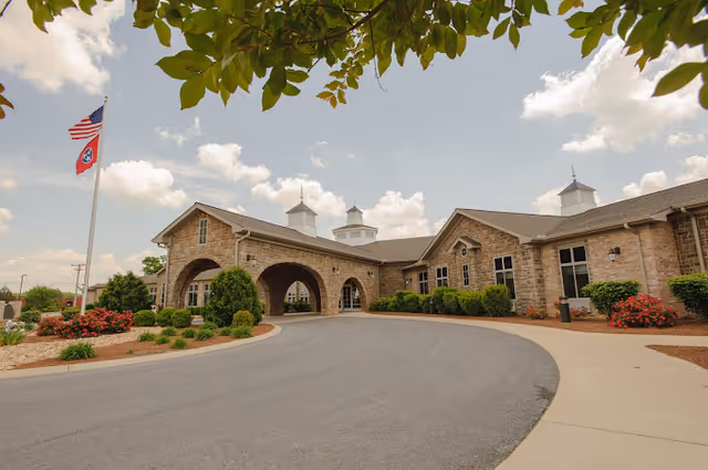 Exterior view of The Pavilion Senior Living at Lebanon, showing a stone building with arched entrance, landscaped bushes and flowers, and two flagpoles with American and Tennessee state flags under a partly cloudy sky.