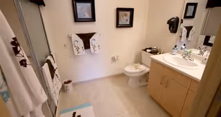 A clean and well-lit bathroom featuring a toilet, a sink with a countertop and cabinet, a mirror above the sink, a towel rack with towels, a shower with a sliding glass door, and decorative framed pictures on the wall.