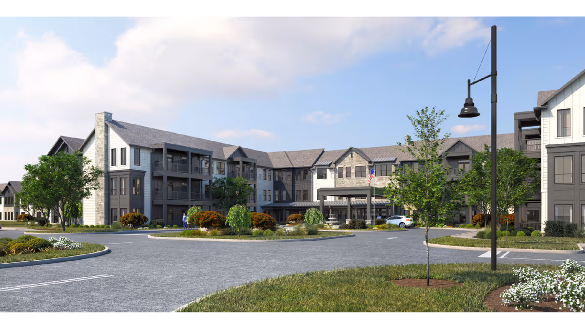 Exterior view of a large, modern senior living facility building with multiple floors, balconies, and a covered entrance. The building is surrounded by landscaped greenery, trees, and a circular driveway with a street lamp and an American flag near the entrance.
