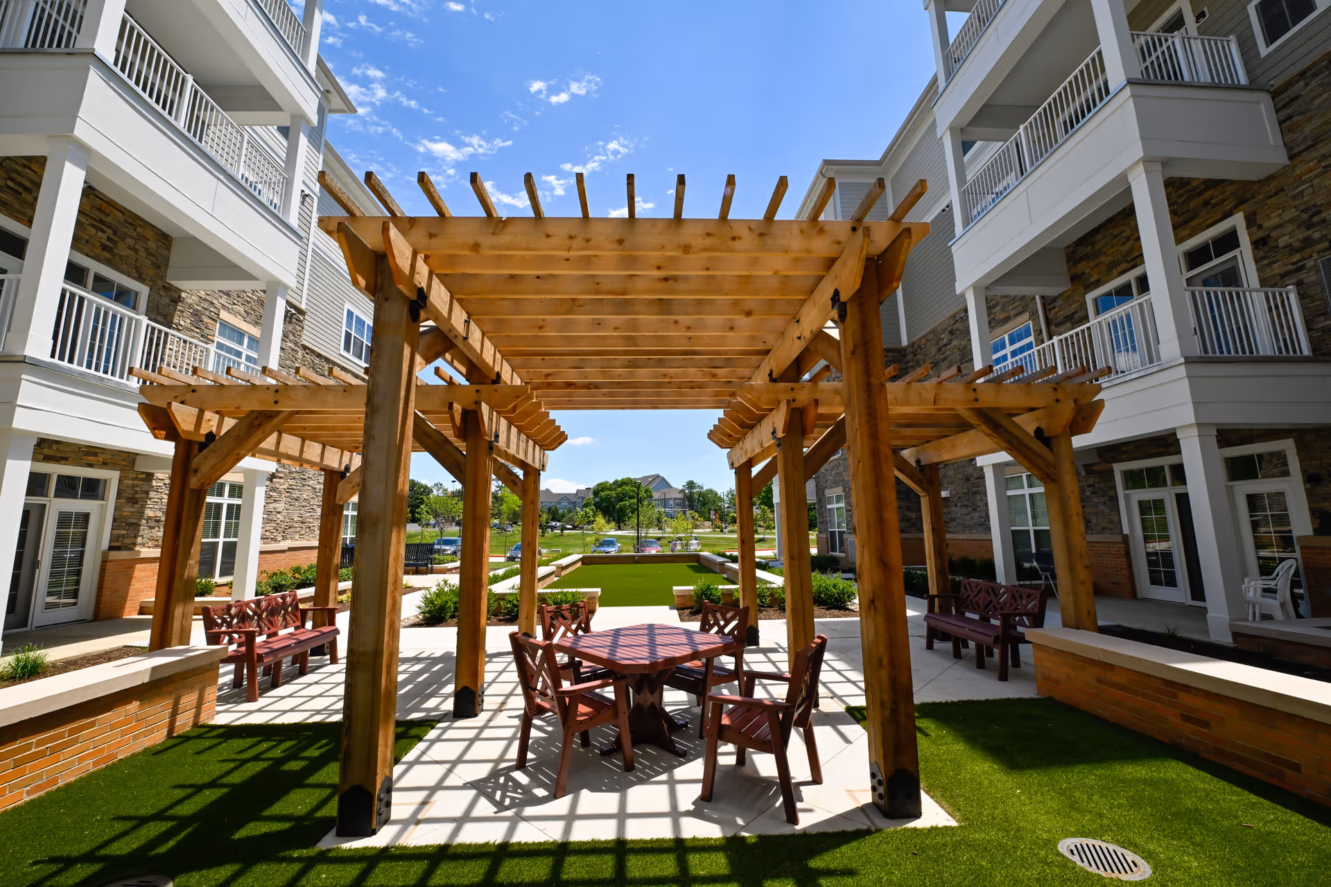 Outdoor courtyard area at The Lumiere of Chesterfield featuring wooden pergolas with seating underneath, surrounded by a multi-story building with balconies and stone accents, under a bright blue sky with some clouds.