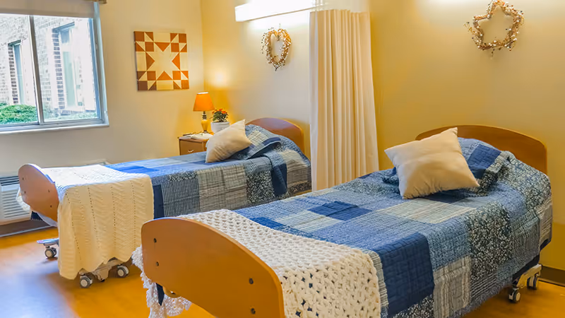 A bright room with two single beds, each covered with blue patchwork quilts and white knitted blankets. Each bed has a pillow and is on wheels. There is a window letting in natural light, a small bedside table with a lamp and a plant, and decorative wall hangings shaped like a star and a heart.