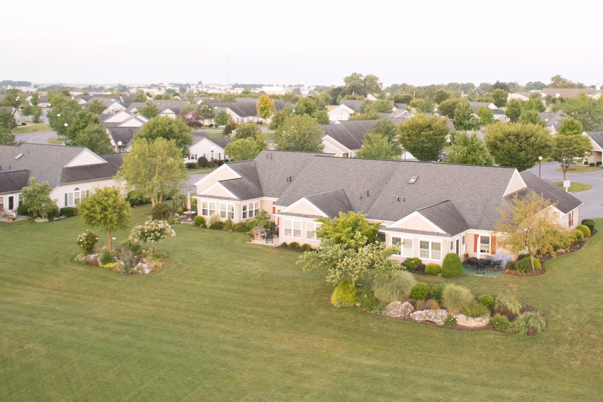 Aerial view of a single-story senior living building surrounded by manicured lawns, landscaped gardens, and nearby houses.