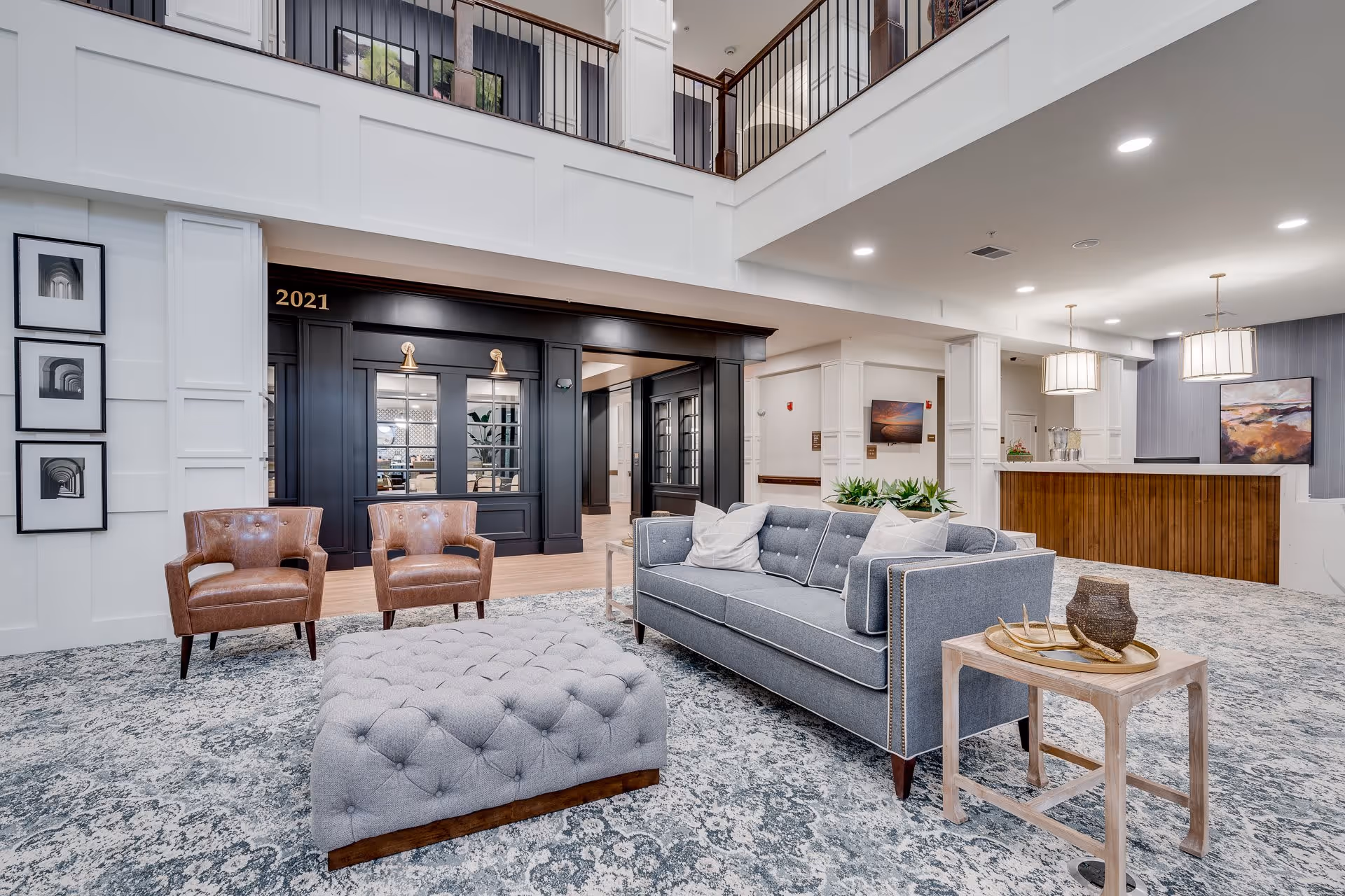A spacious and well-lit senior living facility common area with a gray sofa, two brown leather armchairs, a tufted ottoman, and a small wooden side table with decorative items. The area features a patterned carpet, white paneled walls, and a black framed entrance labeled 2021. There is a reception desk with pendant lights and artwork on the walls in the background.