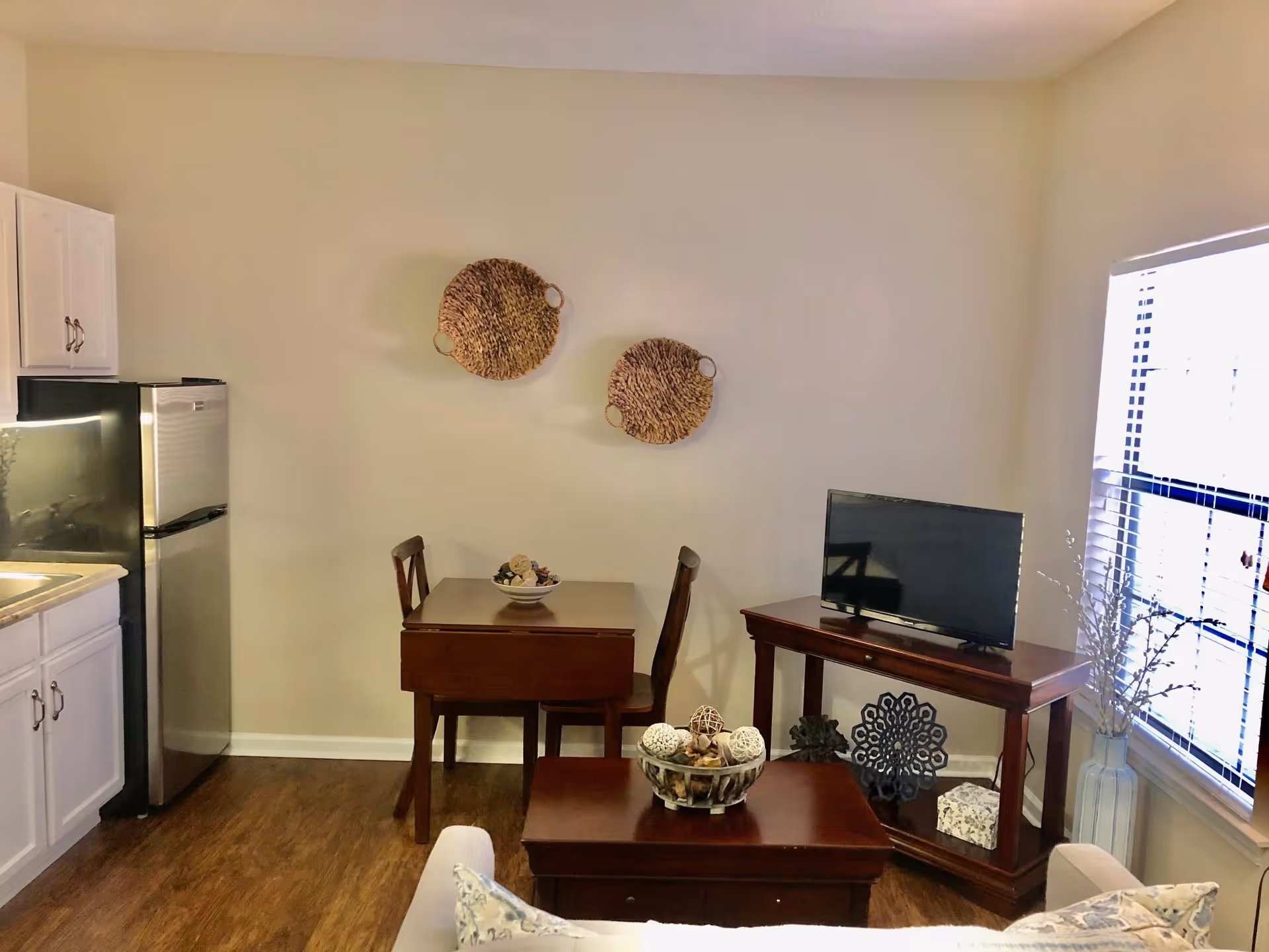 A small living area with a wooden table and two chairs, a flat-screen TV on a wooden stand, and a coffee table with decorative items. The room has light-colored walls with two round woven wall hangings and a window with blinds letting in natural light. Part of a kitchen with white cabinets and a stainless steel refrigerator is visible on the left.
