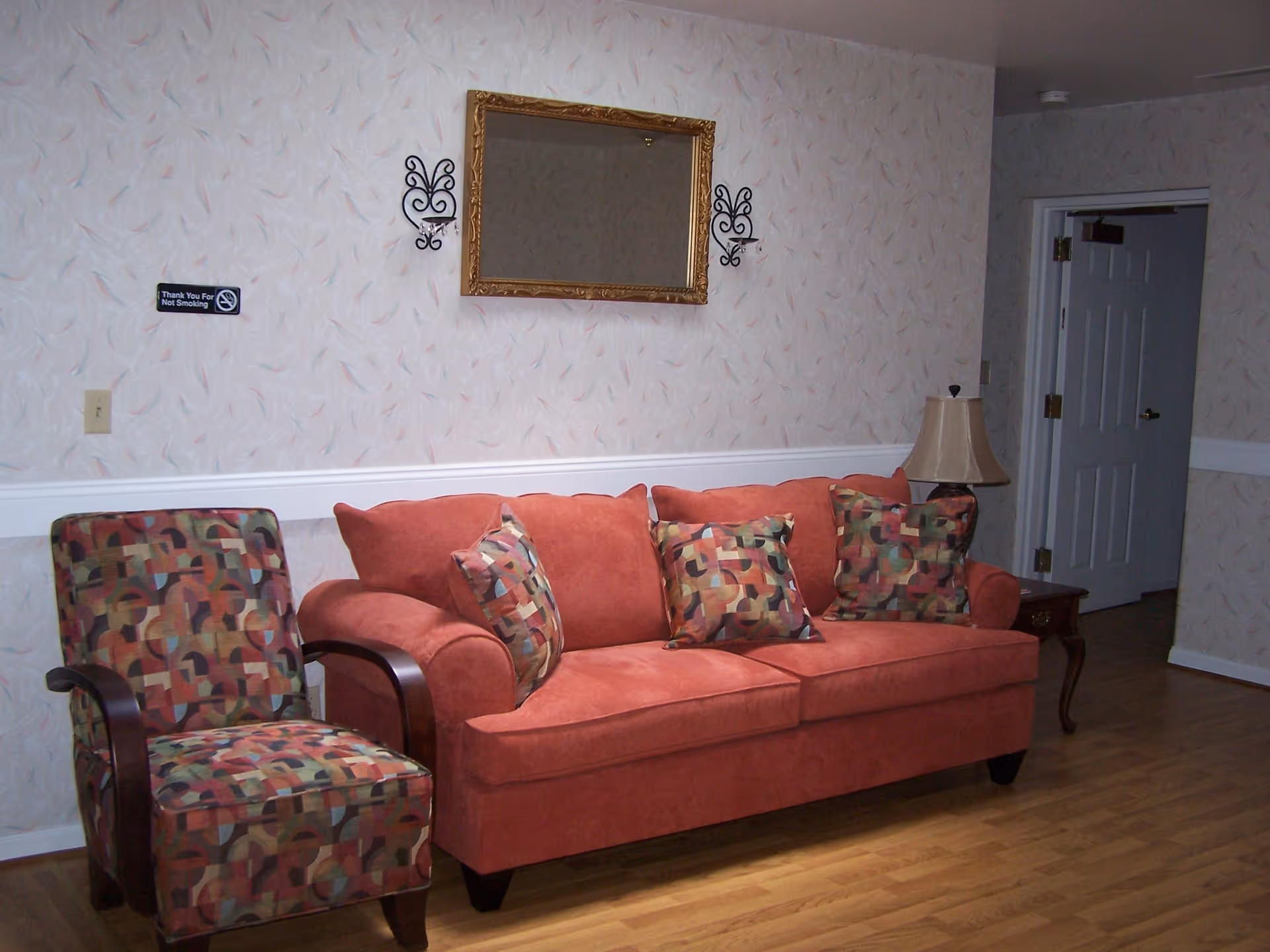 A living room area with a red sofa adorned with three patterned cushions, a matching patterned armchair, a wooden side table with a lamp, a gold-framed mirror on the wall, and a 'Thank You For Not Smoking' sign. The room has light-colored wallpaper with a subtle pattern and wooden flooring.