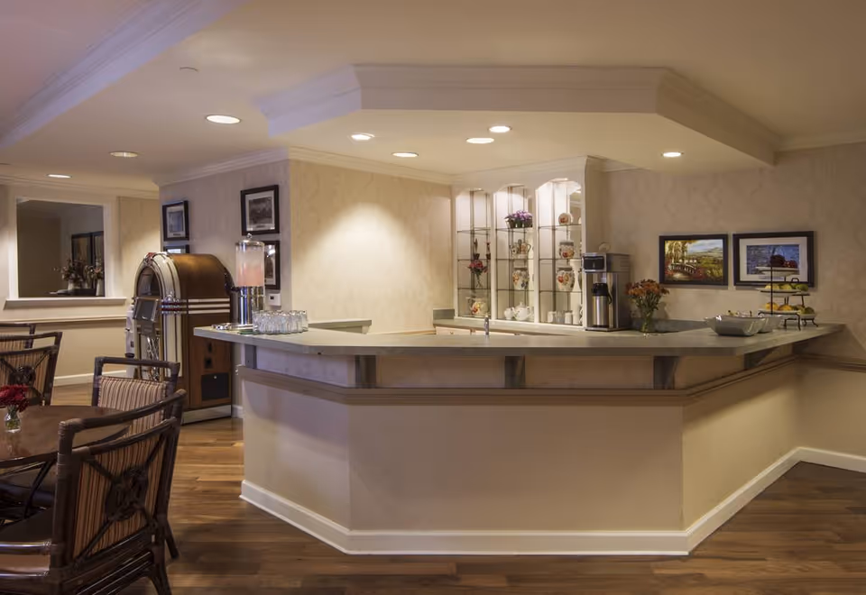 Interior view of a senior living facility's common area featuring a large beige counter with a coffee machine, glassware, and decorative items. There are framed pictures on the wall, a vintage jukebox, and wooden chairs with cushions around tables. The space has warm lighting and wooden flooring.