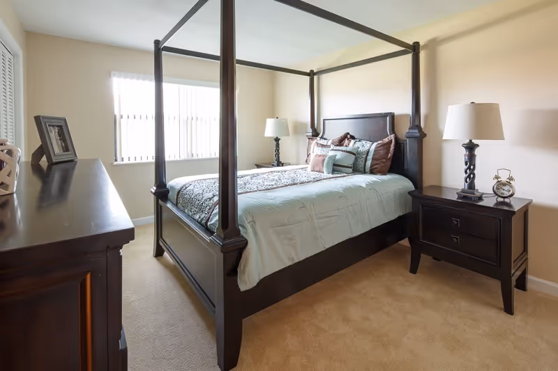 A well-lit bedroom with a large dark wood four-poster bed dressed in light blue and brown bedding. There are two matching dark wood nightstands on either side of the bed, each with a lamp. A dark wood dresser with a framed photo is positioned against the left wall. The room has beige carpet and light-colored walls with a window covered by vertical blinds.