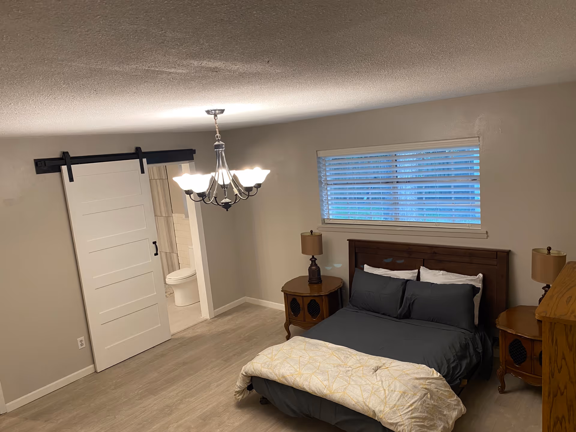 A furnished bedroom with a bed, two nightstands and lamps, a chandelier, a window with blinds, and a sliding barn door leading to a bathroom.