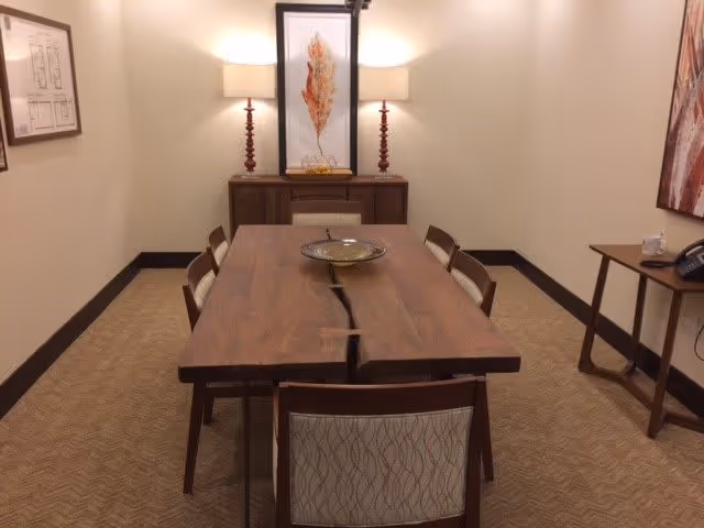 A small conference or meeting room with a wooden table surrounded by six chairs. The table has a natural wood finish with a decorative bowl in the center. At the back of the room, there is a wooden sideboard with two lamps and a framed artwork featuring a leaf. The walls are beige, and there is framed artwork on the left and right walls. The room has carpeted flooring with a subtle pattern.