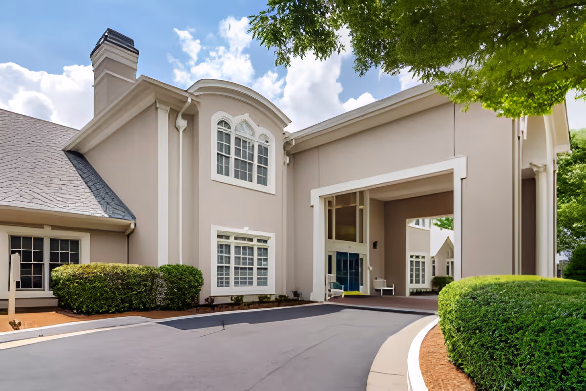 Exterior view of a senior living facility building with beige walls, white trim, multiple windows, a covered entrance, and neatly trimmed bushes along a curved driveway under a partly cloudy sky.