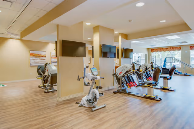 A bright exercise room with stationary and recumbent bikes, mounted TVs, and wood-look flooring.