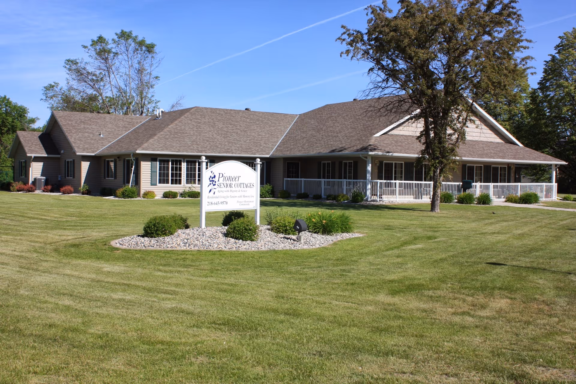 Exterior view of a single-story building with beige siding and a brown roof, surrounded by a well-maintained lawn and landscaping. A white sign in front reads 'Pioneer Senior Cottages' with additional text about the facility. Trees are visible in the background under a clear blue sky.