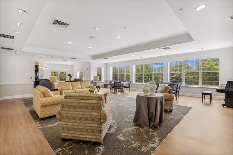 A spacious, well-lit living room area with large windows allowing natural light to fill the space. The room features comfortable patterned armchairs and a sofa arranged around a central coffee table on a decorative rug. In the background, there is a dining area with tables and chairs, and a piano is visible on the right side near the windows.