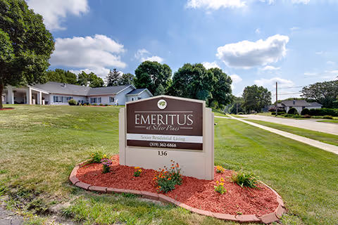 Freestanding sign for 'Emeritus' on a landscaped lawn in front of a single-story senior living building under a blue sky.
