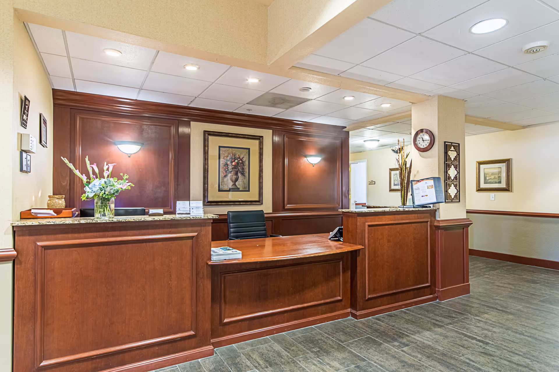 Reception area with a wooden front desk, a black office chair, a vase with flowers, wall sconces, framed artwork, a clock on a pillar, and informational brochures on the desk.