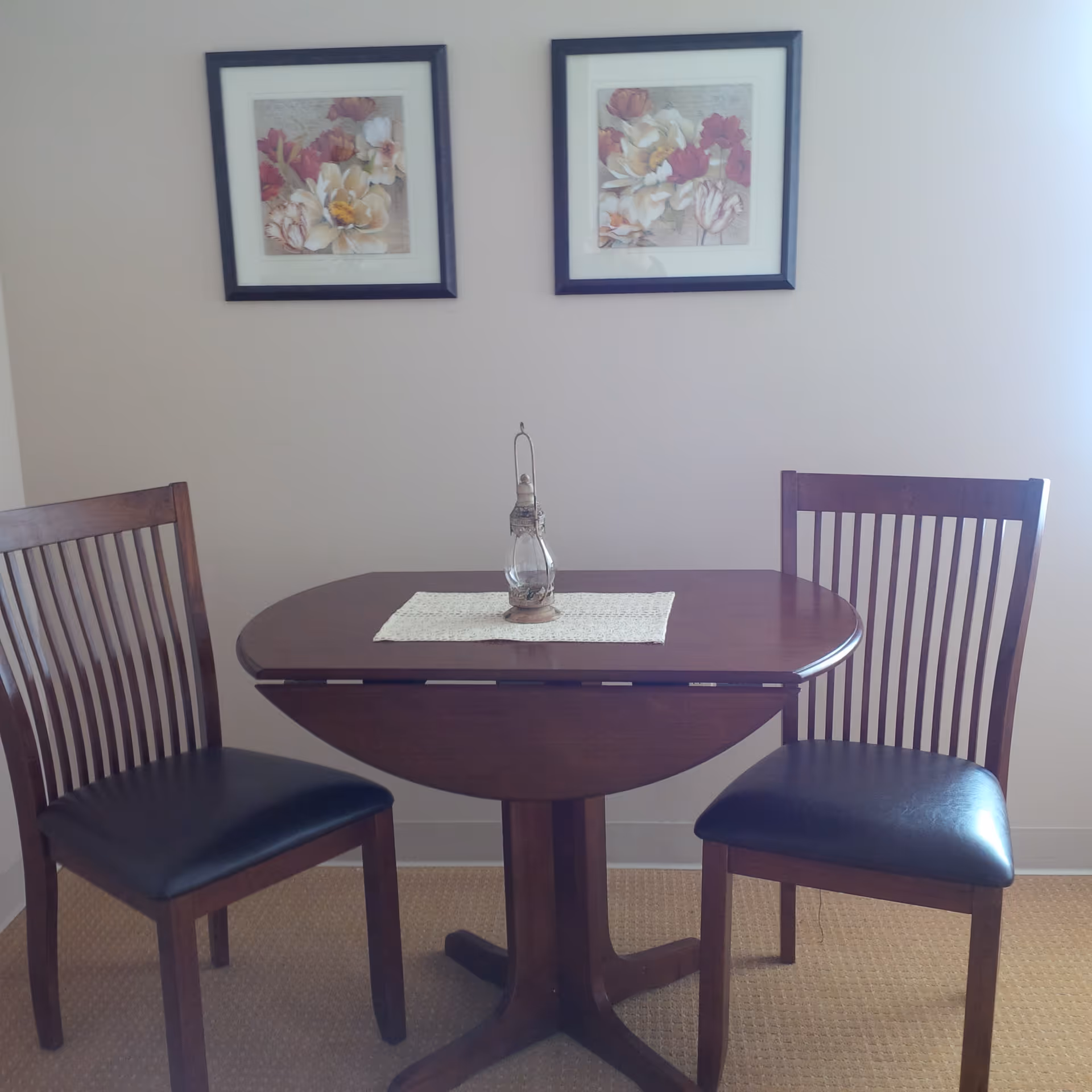 A small wooden dining table with two matching wooden chairs with black cushioned seats. On the table is a white lace runner with a decorative lantern placed in the center. Two framed floral paintings hang on the beige wall behind the table.