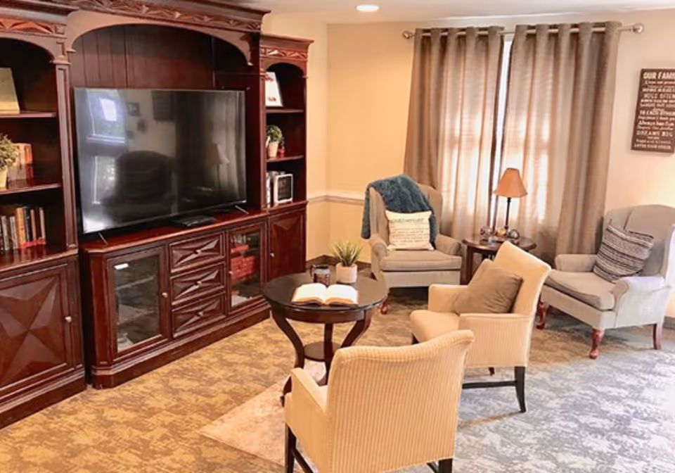 A cozy living room with a large dark wood entertainment center holding a flat-screen TV, surrounded by shelves with books and decorative items. In front of the TV, there is a round wooden coffee table with an open book and a small plant. Four upholstered armchairs are arranged around the coffee table, with a side table and lamp near the window covered by beige curtains.