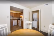 View of a dining area with a round wooden table and white chairs. In the background, there is a kitchen with wooden cabinets, a dishwasher, stove, and refrigerator. To the right, there is a small laundry area with a stacked washer and dryer behind an open door.