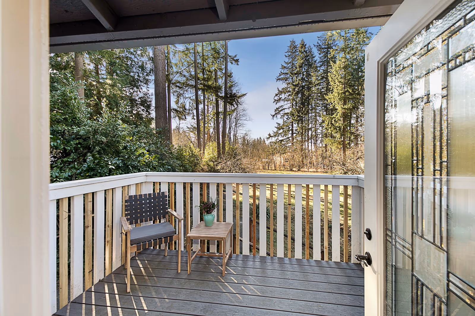 View from an open door onto a small wooden balcony with a single chair and a small table holding a green vase with flowers. The balcony overlooks a wooded area with tall trees and a clear blue sky.