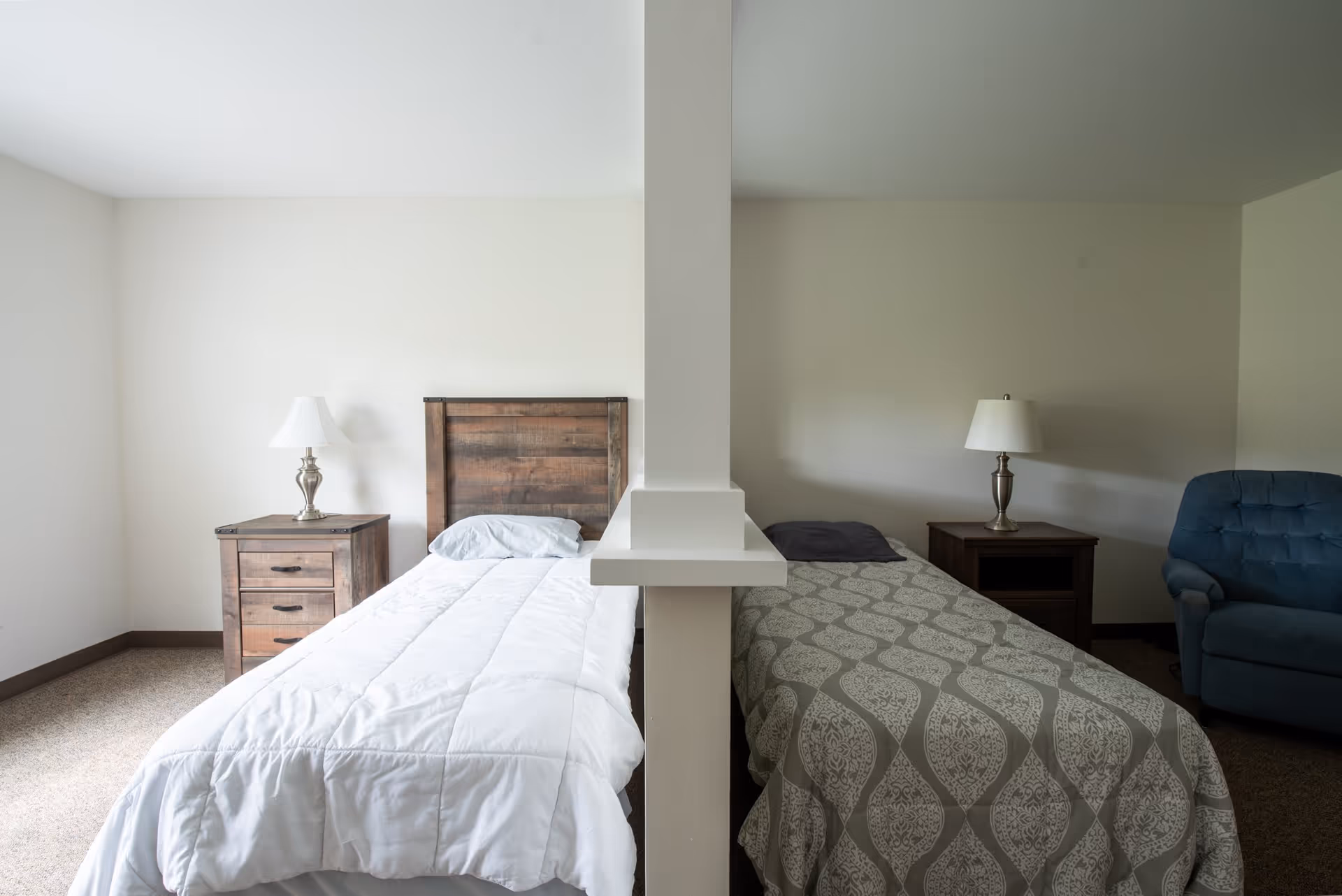 A shared bedroom divided by a partial wall with two single beds on either side. Each bed has a nightstand with a lamp. One bed has a white comforter and light blue pillow, the other has a patterned gray and white comforter with a dark pillow. There is a blue armchair next to the bed on the right side.