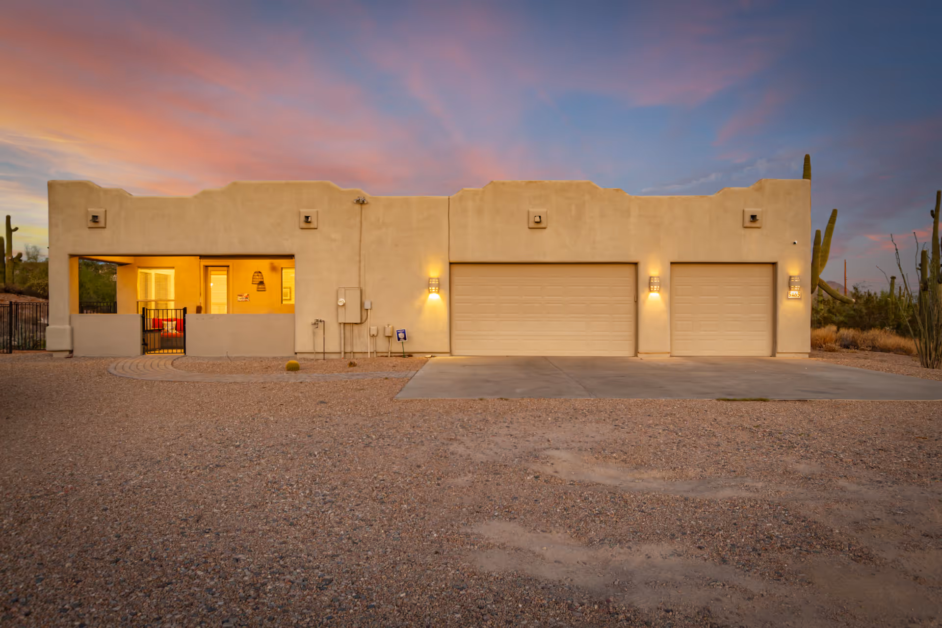 Front exterior of a single-story stucco building with three garage doors and a lit porch at sunset in a desert setting.