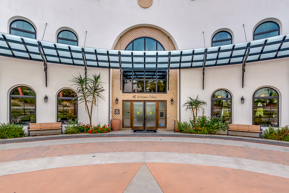Front entrance of a senior living facility with a curved glass canopy above the double doors. The building has arched windows on either side, two benches, potted plants, and landscaped flower beds.